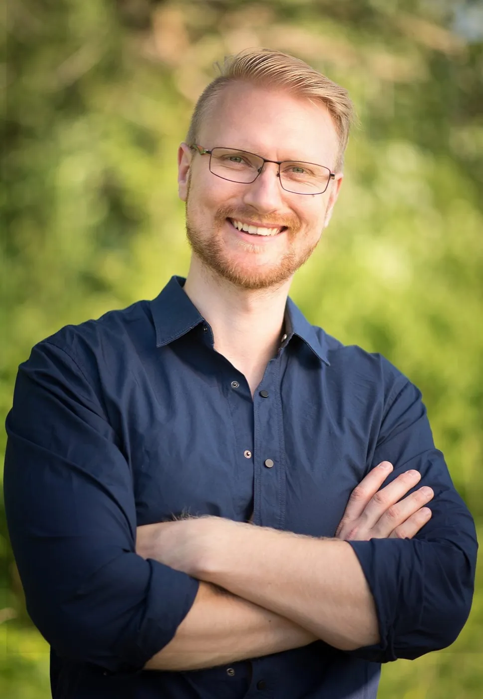 Spas Angelov smiling with his arms crossed, in a navy shirt, trees behind him